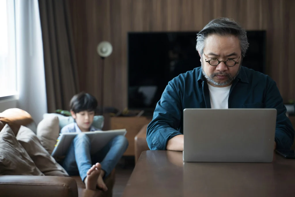 A man learns on a computer while a child draws in the background